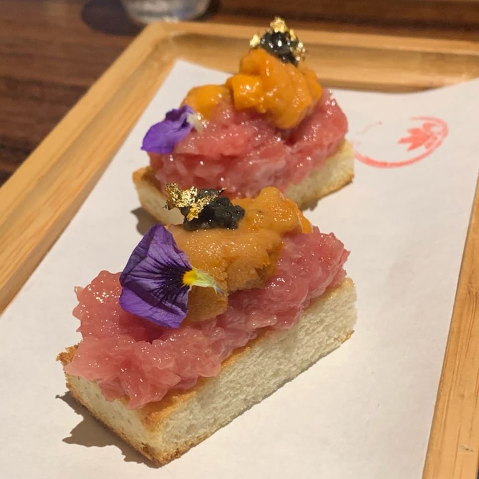Two pieces of tuna tartare on bread, topped with uni, caviar, gold flakes, and purple flowers.
