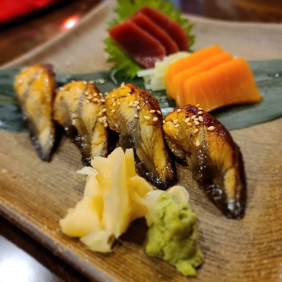 Four pieces of eel nigiri with sesame seeds, ginger, wasabi, tuna sashimi, and orange slices.